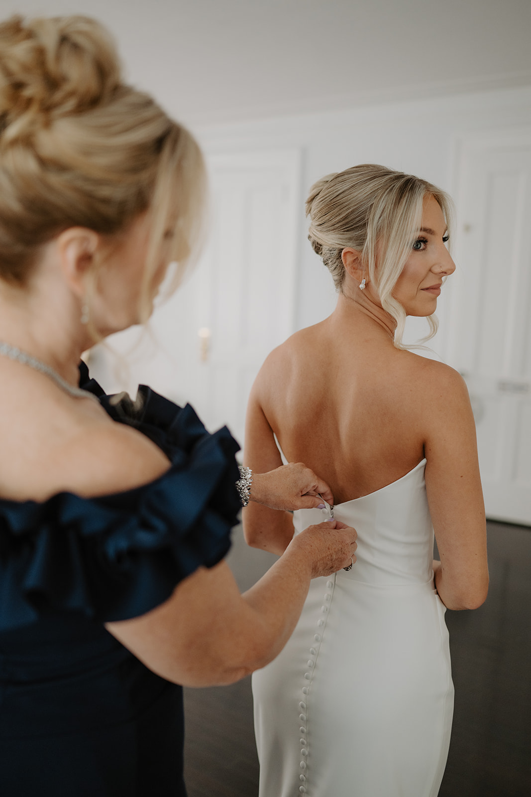 Mother of the bride buttons the wedding gown as the bride looks back over her shoulder, smiling.